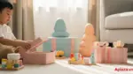 Cảnh bé 3 tuổi mở quà sinh nhật trong phòng khách sáng, xung quanh là đồ chơi cỡ lớn an toàn, bề mặt bo tròn và màu sắc dịu, nhấn mạnh chọn quà đúng độ tuổi và an toàn cho bé.